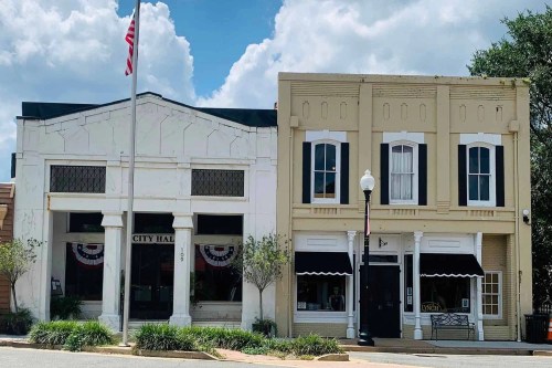 Two historic buildings side by side: a white City Hall with flags and a tan storefront with black awning, under a cloudy sky.