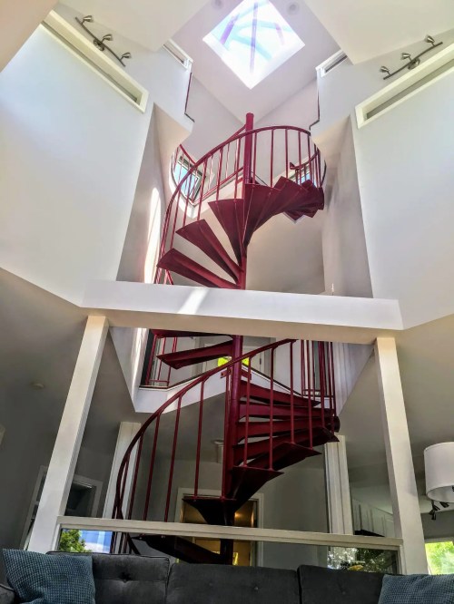 A red spiral staircase ascends through a bright, modern interior with large windows and a skylight above.