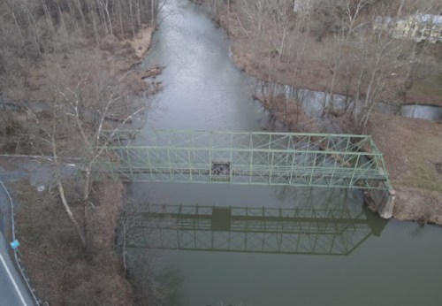 Aerial view of a green metal bridge over a calm river, surrounded by bare trees and a grassy bank.
