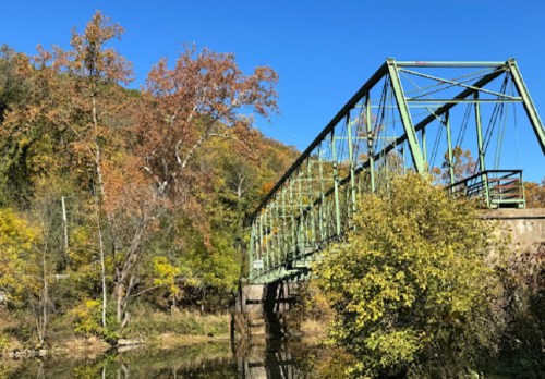 A green metal bridge spans a calm river, surrounded by autumn foliage and clear blue skies.