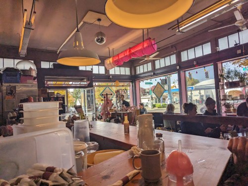 A cozy café interior with a wooden counter, colorful lights, and patrons enjoying their time by the windows.