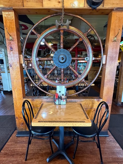 A wooden table with black chairs in a restaurant, framed by a large ship's wheel in the background.