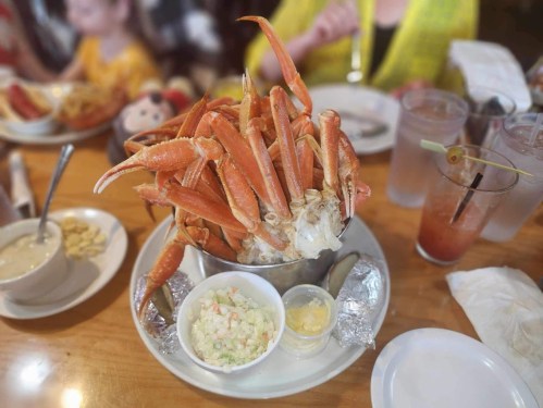 A bucket filled with crab legs, accompanied by coleslaw and dipping sauces, on a wooden table.