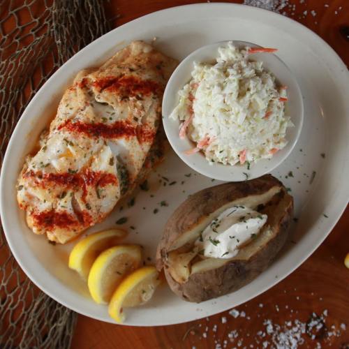 A plate featuring grilled fish with lemon, coleslaw, and a baked potato with sour cream, garnished with herbs.