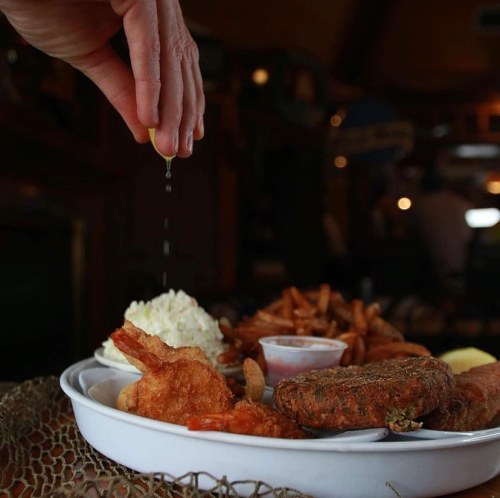 A hand squeezing lemon over a plate of fried shrimp, hush puppies, fries, and coleslaw in a cozy restaurant setting.