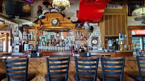 A rustic bar with wooden shelves, various bottles, and a clock, featuring empty black chairs in front.