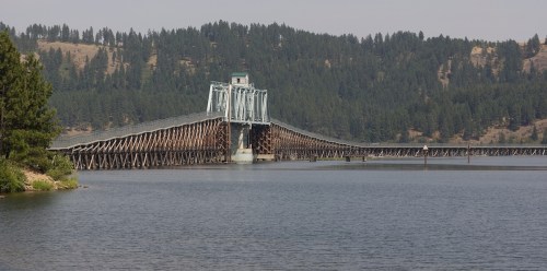 A large wooden bridge spans a calm lake, surrounded by dense green forests and distant hills.