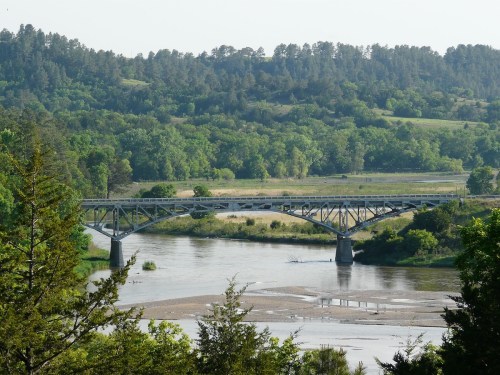 A metal bridge spans a river, surrounded by lush green trees and hills under a clear sky.