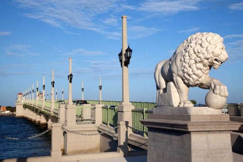 A stone lion statue stands beside a green bridge with lampposts, overlooking a calm body of water under a blue sky.