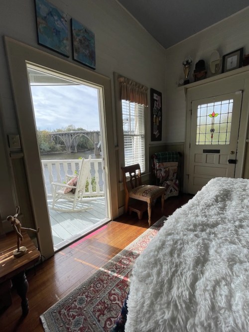 Cozy room with a view of a bridge, featuring a chair, a bed with a fluffy blanket, and sunlight streaming in.