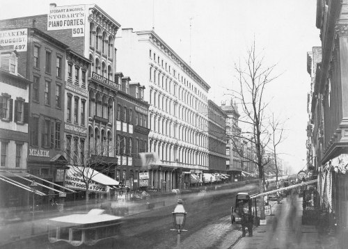 Historic black-and-white street scene featuring buildings, storefronts, and a horse-drawn carriage on a busy city street.