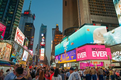 Crowd gathered in Times Square, vibrant billboards illuminated at dusk, showcasing colorful advertisements and city life.