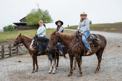 Three women on horseback, smiling, with a rustic barn in the background on a cloudy day.