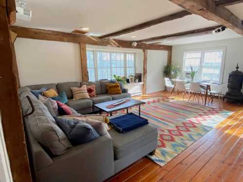 Cozy living room with a gray sectional sofa, colorful rug, wooden beams, and a dining area with natural light.