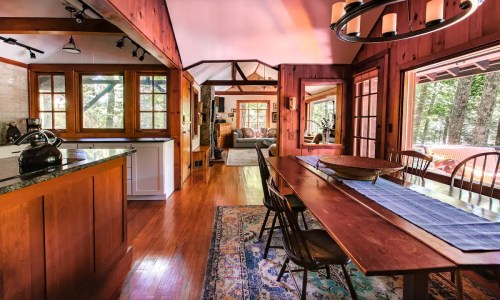 Cozy wooden interior featuring a dining table, kitchen, and living area with large windows and natural light.