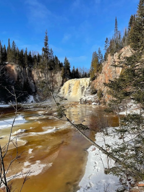 A waterfall cascades over rocky cliffs, surrounded by snow and evergreen trees under a clear blue sky.
