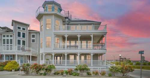 A large, multi-level beach house with a wraparound porch, surrounded by colorful flowers under a pink sunset sky.