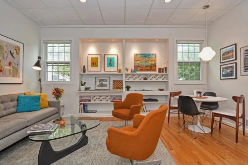 A modern living room with a gray sofa, orange chairs, a glass coffee table, and colorful artwork on the walls.