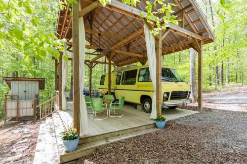 A yellow vintage RV parked under a wooden gazebo surrounded by greenery, with a table and chairs on the deck.