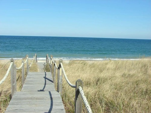 A wooden walkway leads to a serene beach with gentle waves and tall grass under a clear blue sky.