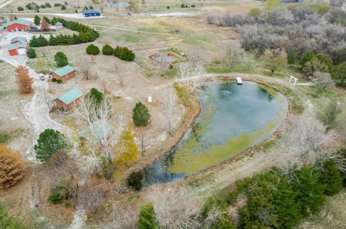 Aerial view of a rural landscape featuring a pond, trees, and several small buildings in a grassy area.