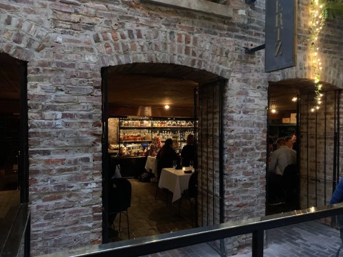 A cozy restaurant with brick walls, featuring tables and a well-stocked bar visible through an open entrance.