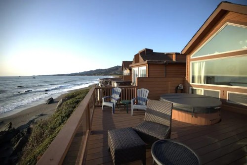 Cozy deck overlooking the ocean, featuring chairs and a hot tub, with a clear sky and gentle waves in the background.