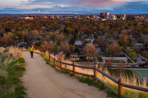A person walks along a dirt path overlooking a cityscape with trees in autumn colors and buildings in the distance.