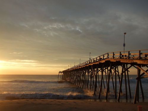 A wooden pier extends into the ocean at sunset, with gentle waves and a cloudy sky in the background.