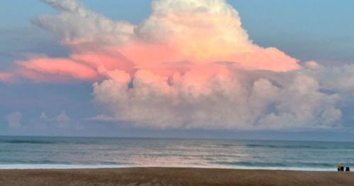 A serene beach scene at sunset, featuring soft pink clouds above calm ocean waters.