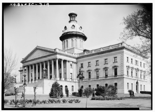 Historic building with a dome and columns, surrounded by trees and gardens, featuring people in the foreground.