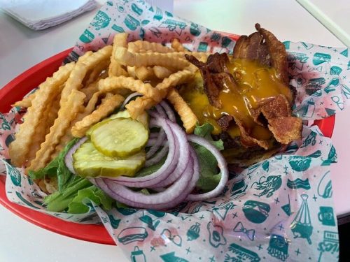A plate with a burger topped with cheese, bacon, lettuce, pickles, and red onion, accompanied by crinkle-cut fries.