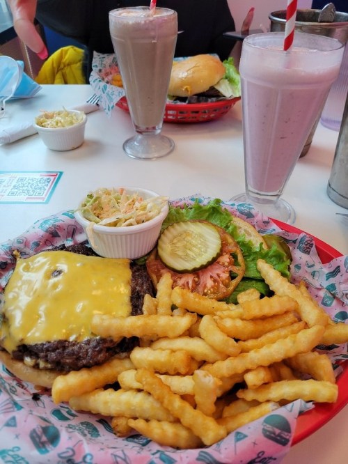 A cheeseburger with fries, coleslaw, and pickles, alongside milkshakes in a diner setting.