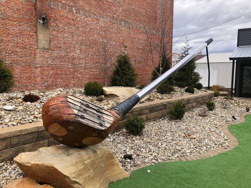 A large, artistic golf club sculpture rests on a rock in a landscaped area with pebbles and small shrubs.
