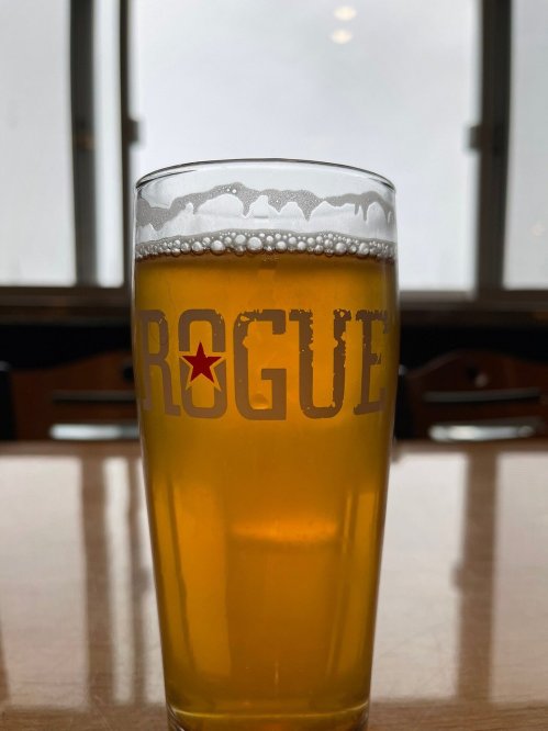 A glass of beer with a frothy top, featuring the word "ROGUE" printed on the side, set against a blurred background.