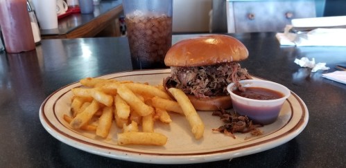 A plate with a pulled pork sandwich, fries, and a small cup of barbecue sauce, alongside a drink.