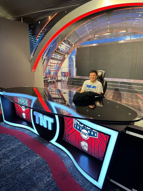 A person sitting at a modern TV studio desk with "TNT" branding, smiling in front of a colorful backdrop.