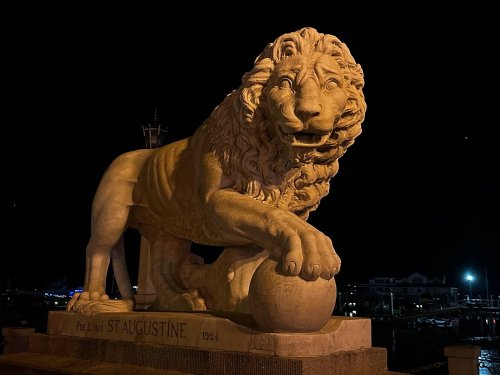A large stone lion statue with a ball, illuminated at night, located in St. Augustine, Florida.