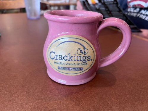 A pink coffee mug with the logo of "Crackings" restaurant, featuring the text "breakfast, brunch, & lunch."