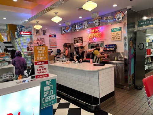 A retro diner counter with staff preparing food, neon signs, and a colorful interior. Customers wait to be seated.