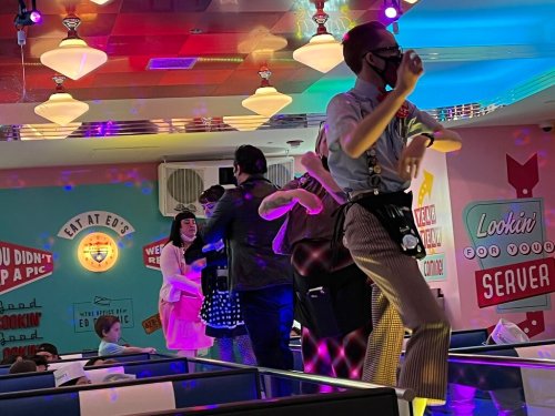 A lively diner scene with people dancing and colorful lights, featuring a retro-themed decor and playful atmosphere.