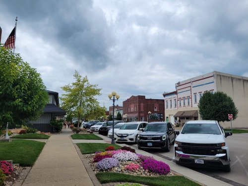 A street lined with parked cars and colorful flower beds, with buildings and cloudy skies in the background.