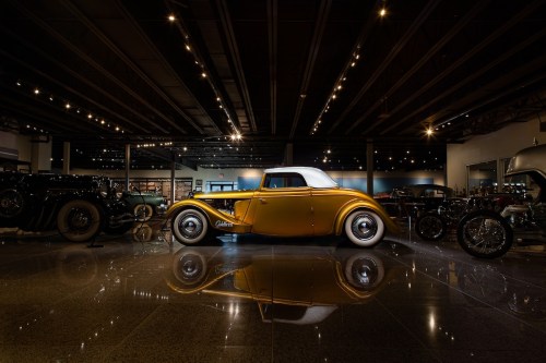 A vintage yellow car with a white top displayed in a well-lit showroom, reflecting on the polished floor.