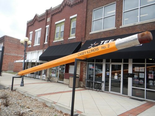 Giant orange pencil sculpture outside a storefront in Casey, Illinois, with brick buildings in the background.