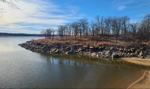 A serene lakeside view with rocky shorelines, trees, and a clear blue sky reflecting on the water.