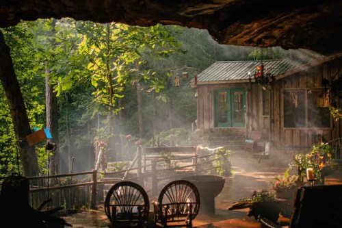 A cozy cabin nestled in a forest, surrounded by greenery and soft morning mist, viewed from a rocky overhang.