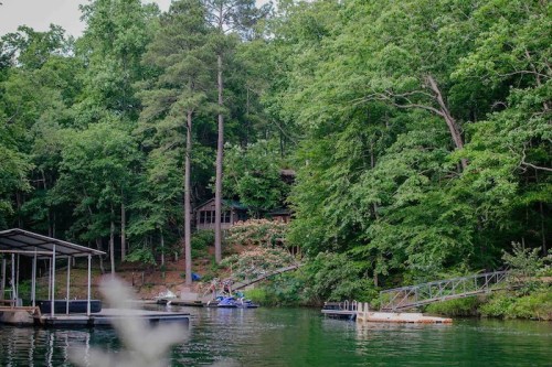 A serene lake scene surrounded by lush trees, featuring a dock and watercraft near a cabin in the background.
