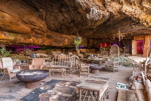 A rustic cave interior with wooden furniture, a fire pit, and plants, illuminated by colorful lights.