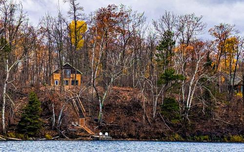 A cozy cabin on a hillside surrounded by autumn trees, overlooking a calm lake.