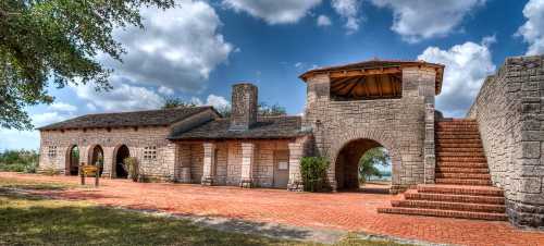 Historic stone building with arches, a staircase, and a scenic sky in the background. Greenery surrounds the area.